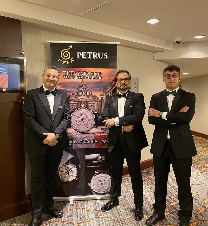 Three men in tuxedos beside a KEFA Petrus banner